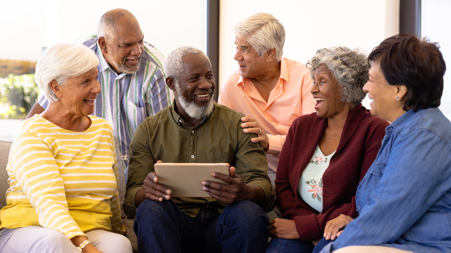 Multiracial senior man holding digital tablet looking at laughing friends while sitting on sofa